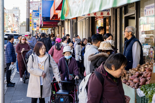 A bustling street scene shows people shopping and walking along a crowded sidewalk with various storefronts. Some wear masks, and a man pushes a stroller.